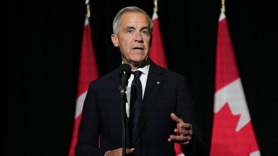 Mark Carney, in a black suit and tie gestures as he stands in front of a microphone. Behind him, against a dark background, are three red and white Canadian flags in a row.