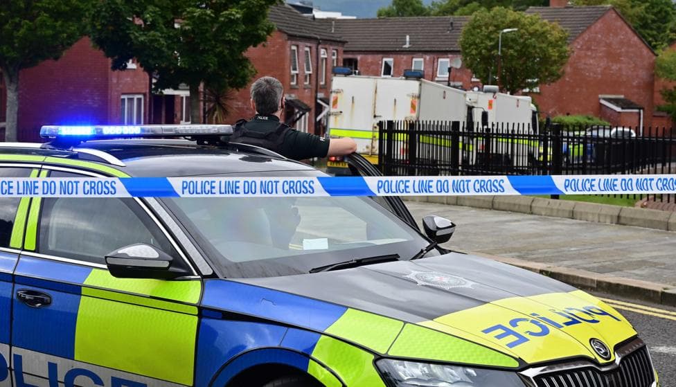 A police officer stands with his back the camera in front of a blue and green liveried police car and police tape which says 'police line do not cross' while some metres away are police bomb disposal vans outside houses in a housing development.