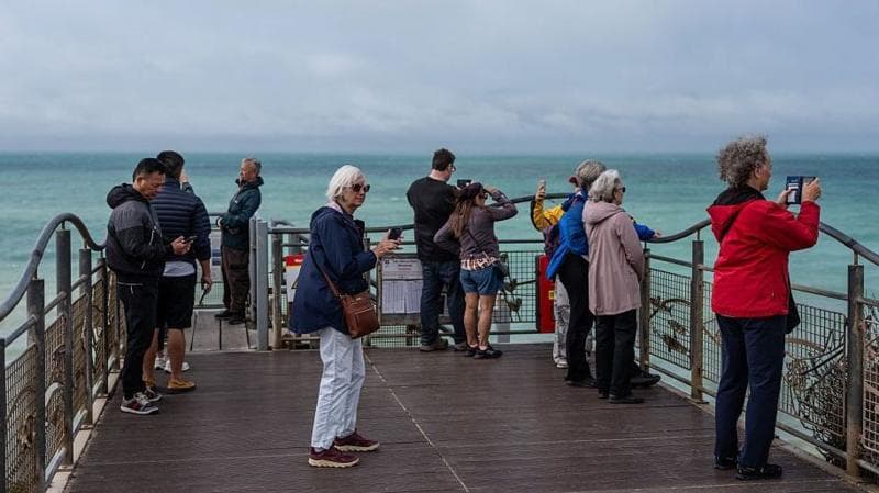 Group of people standing on a pier looking out to sea. Many are wearing coats, and it also looks quite breezy