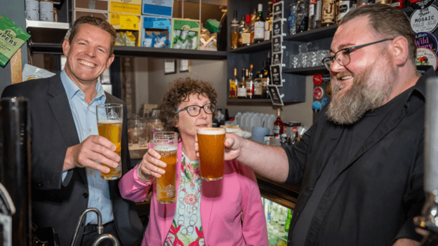 Rhun ap Iorwerth holding a pint and smiling at the camera. He has a dark grey suit and light blue shirt. Next to him is a woman with curly, brown hair and a pink blazer, with a light blue floral top underneath, she too holds a pint. Next to her is a man who smiles with a beard and glasses, and a black shirt, who also holds a pint. The three are stood behind a bar.