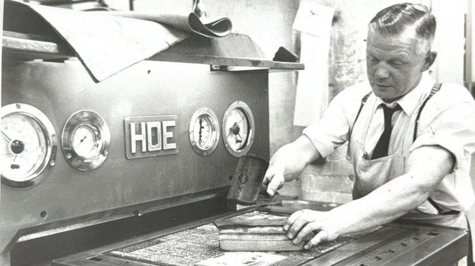 Joseph Paton, a man holding a printer's block in front of an HDE machine. He is wearing a shirt, an apron and a tie.