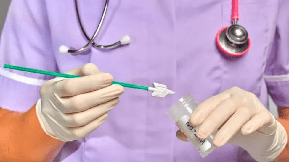 A medic, pictured from the shoulders to waist wearing purple scrubs, with a stethoscope around their neck, wearing white gloves and holding a sample tube.