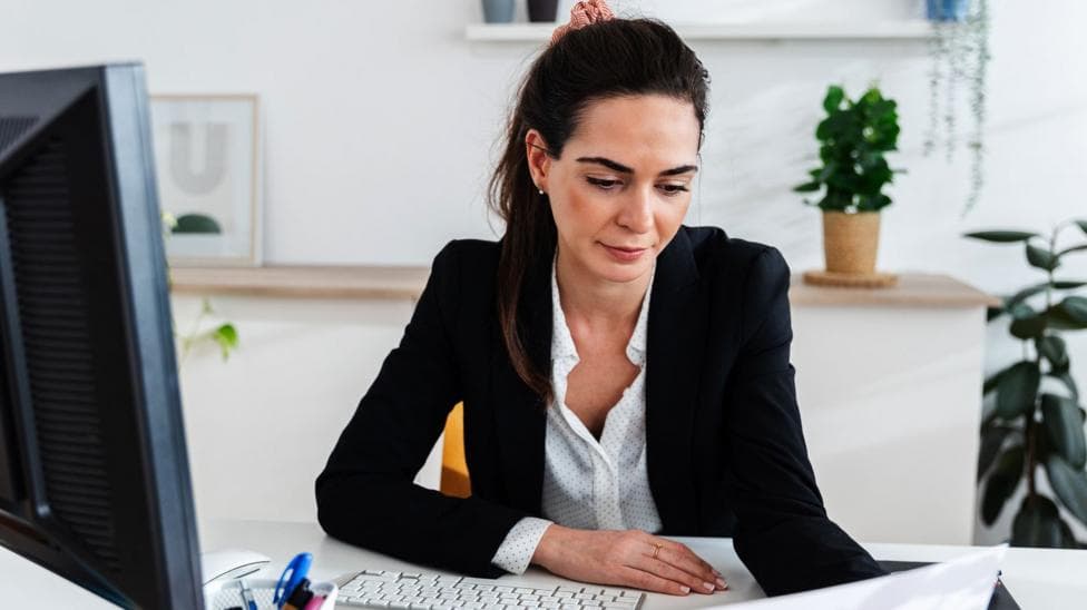 A stock image of a woman with brown hair tied back. She is holding and looking at a page. She is sitting at a desk with a computer and keyboard and is wearing a black blazer.