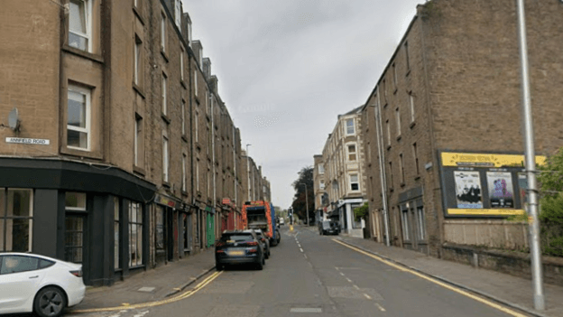 A residential road with cars parked on the left hand side. There are tenements on either side of the road