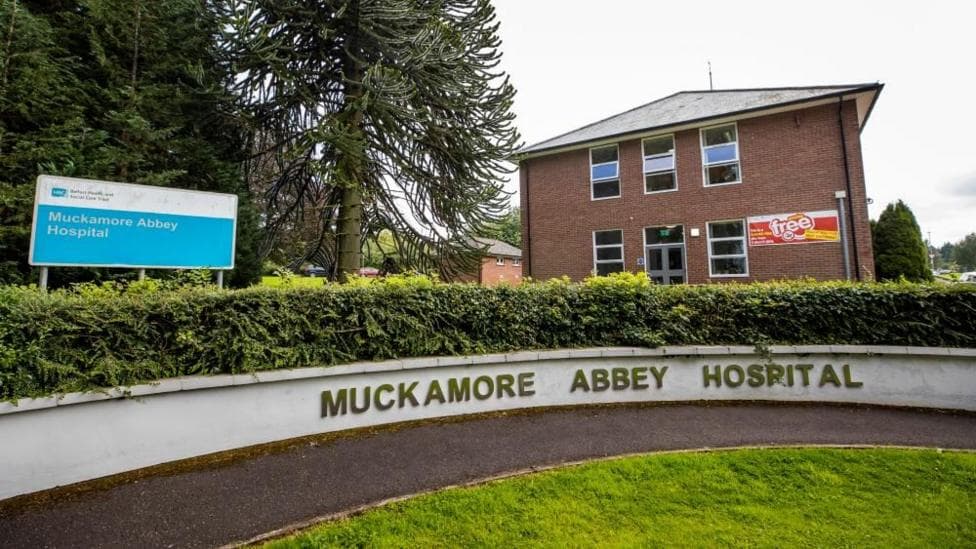 A blue hospital sign that says Muckamore Abbey Hospital on a blue background in white writing. A red brick building is in the background.