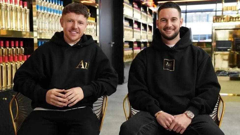 Two men in black hoodies smile at the camera, with gold-coloured bottles of vodka displayed behind them