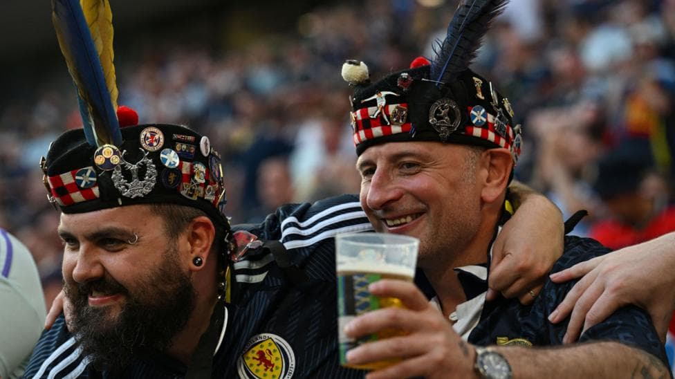 Two Scotland football fans with arms around each other and wearing replica dark blue Scotland tops and bonnets with various lapel pins on it. One of the men has a pint glass with beer in it in one hand.
