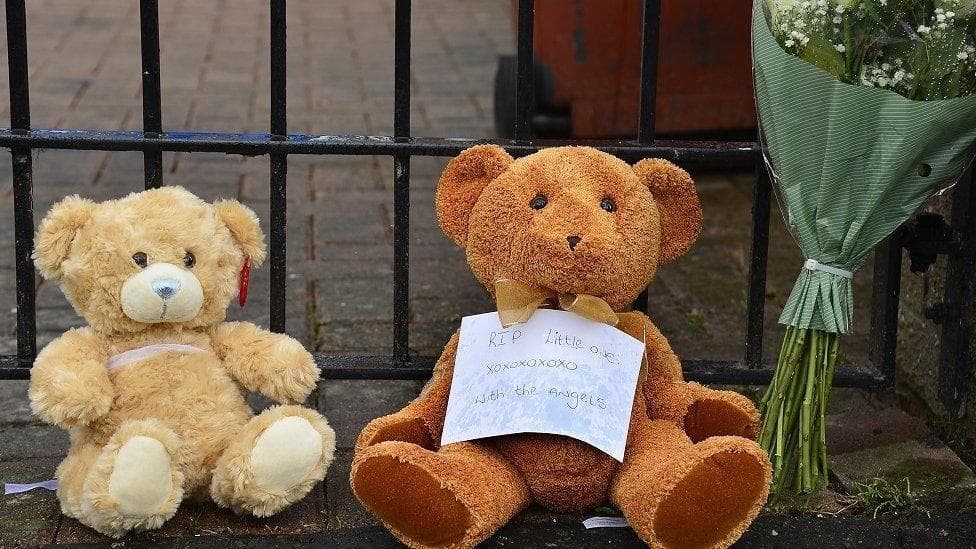 Two teddy bears, small brown furry toys, with a bunch of flowers, against a railing. There is a handwritten note that reads "RIP little one xoxoxoxox with the angels".