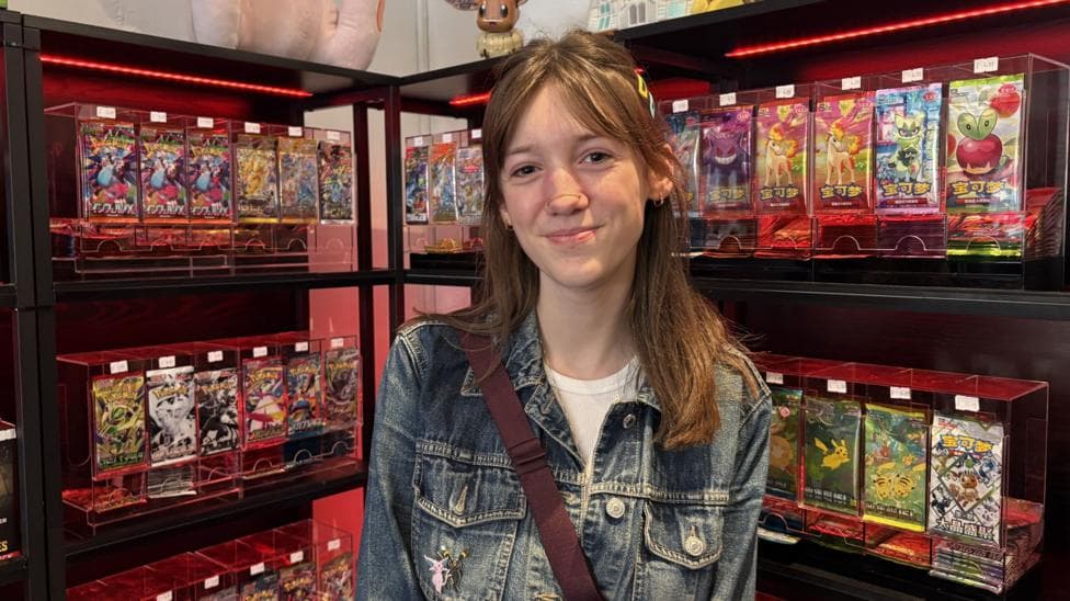 A young woman with dark hair, wearing a denim jacket with a Pokémon badge on it is smiling at the camera. She is standing in front of a display of Pokémon cards in a shop.