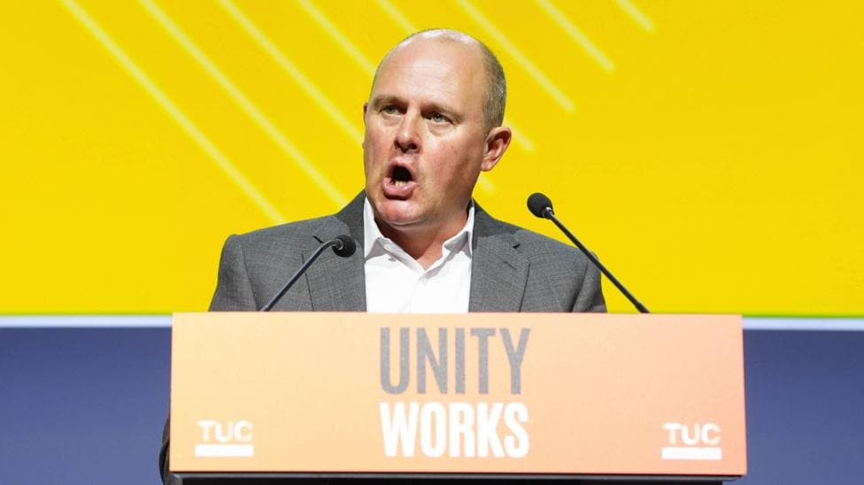 TUC general secretary Paul Nowak speaking at a lectern which has the words "Unity Works" printed on the front. Paul Nowak is wearing a grey suit and a white shirt.