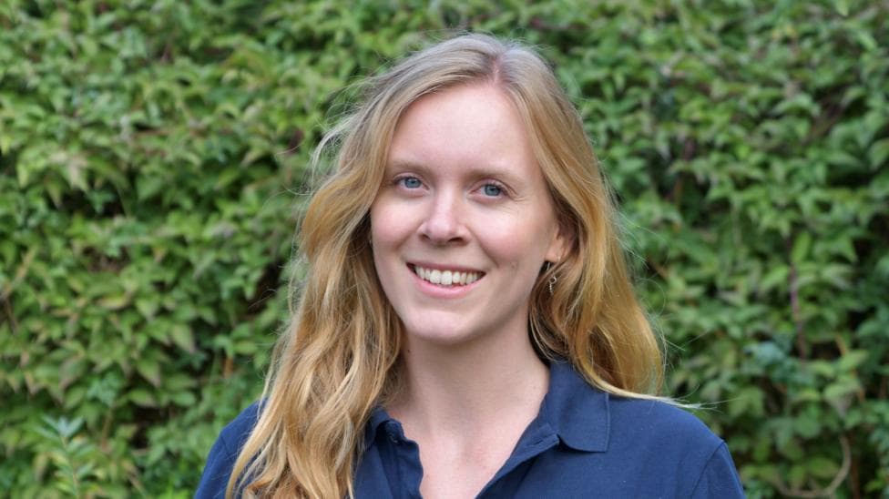 A woman wearing a dark blue polo shirt while standing outside with green bushes behind her.