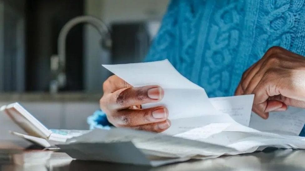 Close up shot of a person looking at bills and receipts on a table next to a calculator in their kitchen. They are wearing a blue jumper and the kitchen sink is in the background.