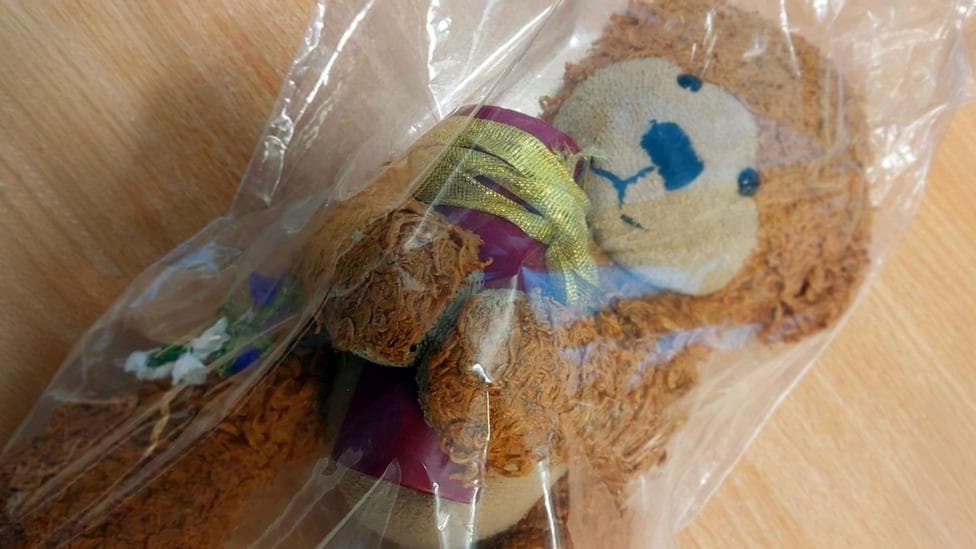 A clear plastic bag containing a brown teddy bear clutching a red urn with a gold ribbon tied round the top. The bag is resting on a light wood table.