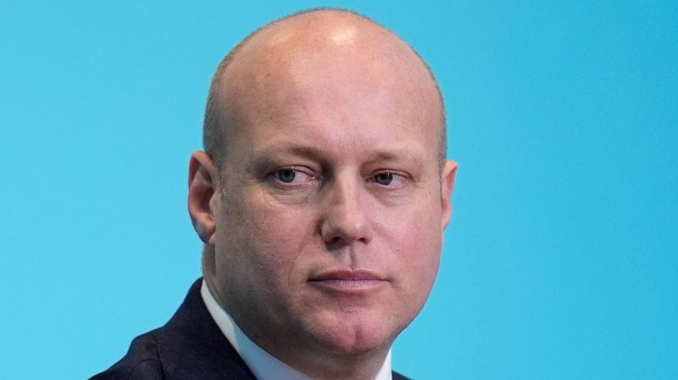 Dan Thomas looking off to the left hand side of the camera, with a blue background behind him. He is wearing a suit and white shirt.