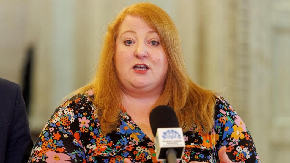 Naomi Long speaking into a microphone. She is wearing a patterned top and has long red hair.