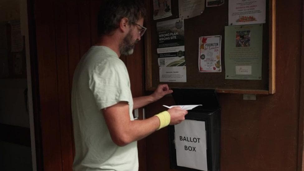 A man in a green t-shirt puts his ballot paper into a black ballot box.
