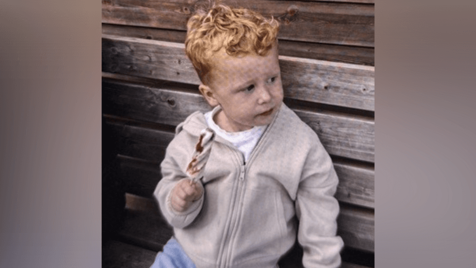 A boy with curly ginger hair, sitting on a wooden bench, wearing a cream-coloured hoodie, and blue denim jeans. He appears to be holding an iced lolly, and is looking to the right of the frame.