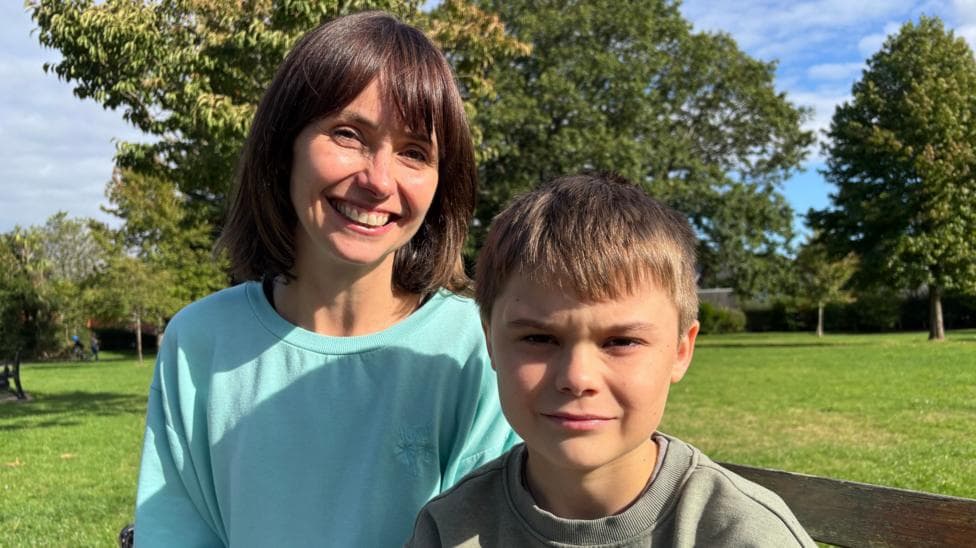 A woman and a child sit in the park. They both look at the camera - it is a head and shoulders shot. The woman smiles, and has a dark brown bob. She has a turquoise jumper on. The boy has short light-brown hair and a light green jumper on.