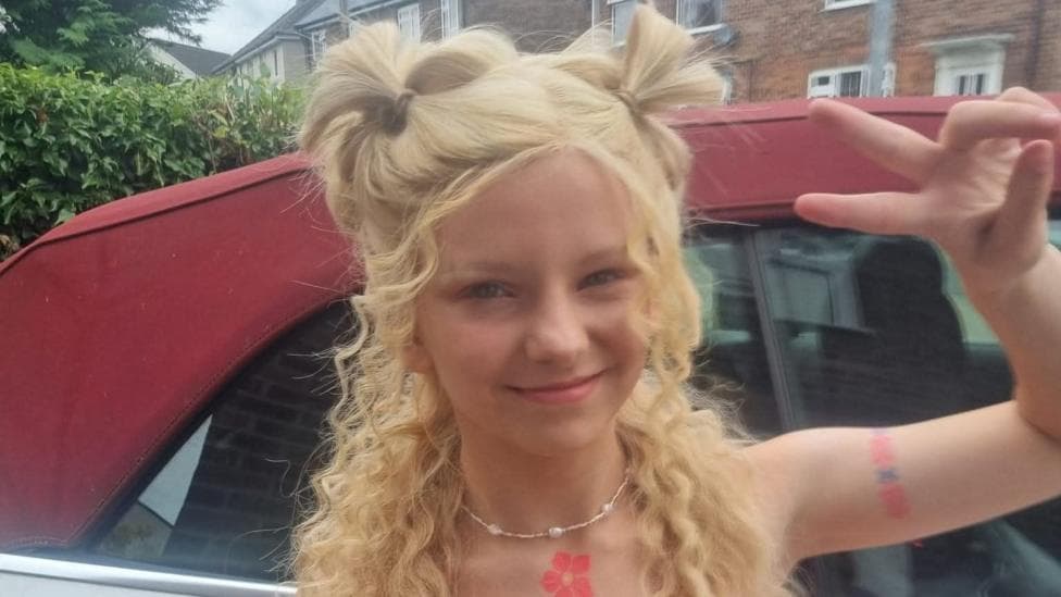 Kizzi-Rae smiles at the camera, she is wearing a blonde wig which is styled half up-half down, there is long curly hair over her shoulders and half of it is tied up in buns on the top of her hair. She is making a peace sign at the camera. She is wearing a necklace and is stood in front of a red car