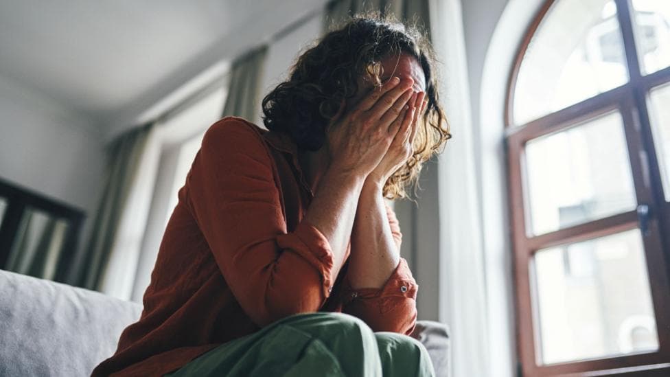 A stock image of a woman sitting on a sofa with her hands covering her face. She is wearing an orange top and green trousers. Her brown wavy hair comes down to her chin. Her body language shows she is very upset. Behind her is a large window letting in daylight.