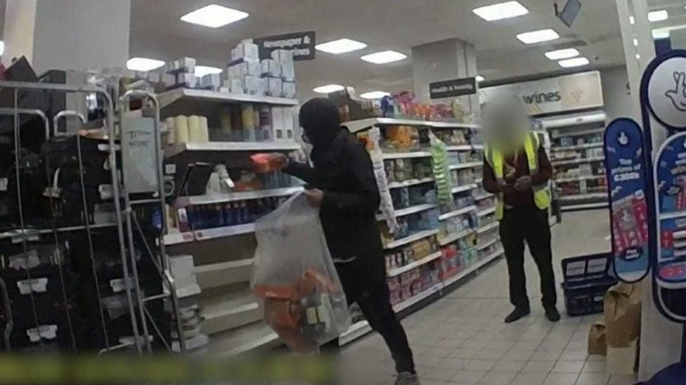CCTV screenshot of a shoplifter in the act in a store aisle. The shoplifter is wearing a black hoodie and is in the process of filling a large transparent bag with goods, while a shopkeeper in high vis stands by.