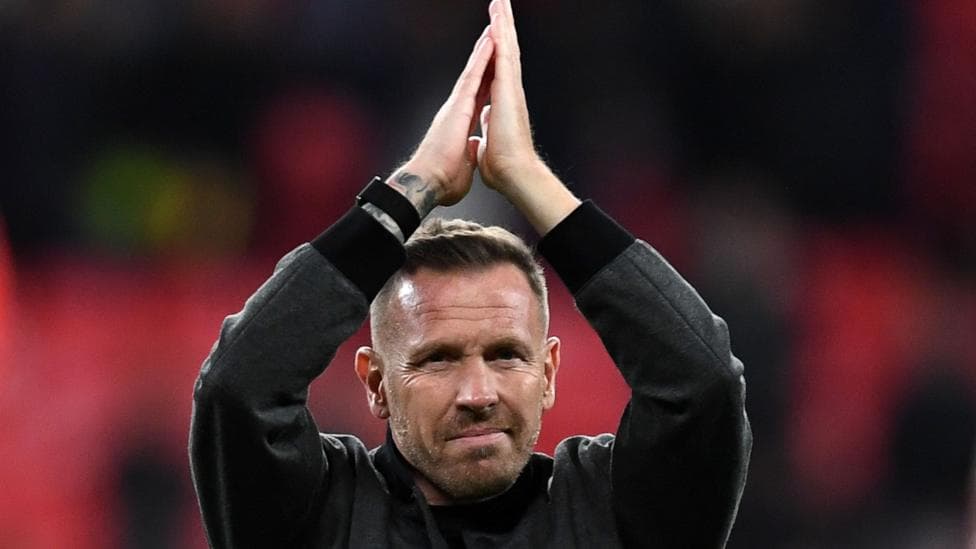 Craig Bellamy in a black jacket faces camera and claps hands above his head towards fans
