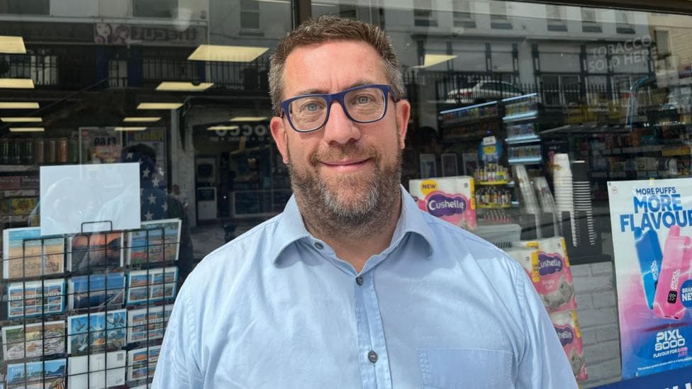 Andrew Bond is a man wearing a blue shirt and blue glasses. He is standing outside a store with a shelf of pictures and post cards behind him.