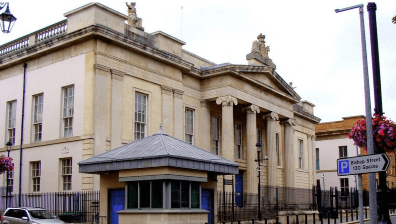 Shows a large court building with four pillars at the front, a security hut to the left and a silver-coloured car to the extreme left.