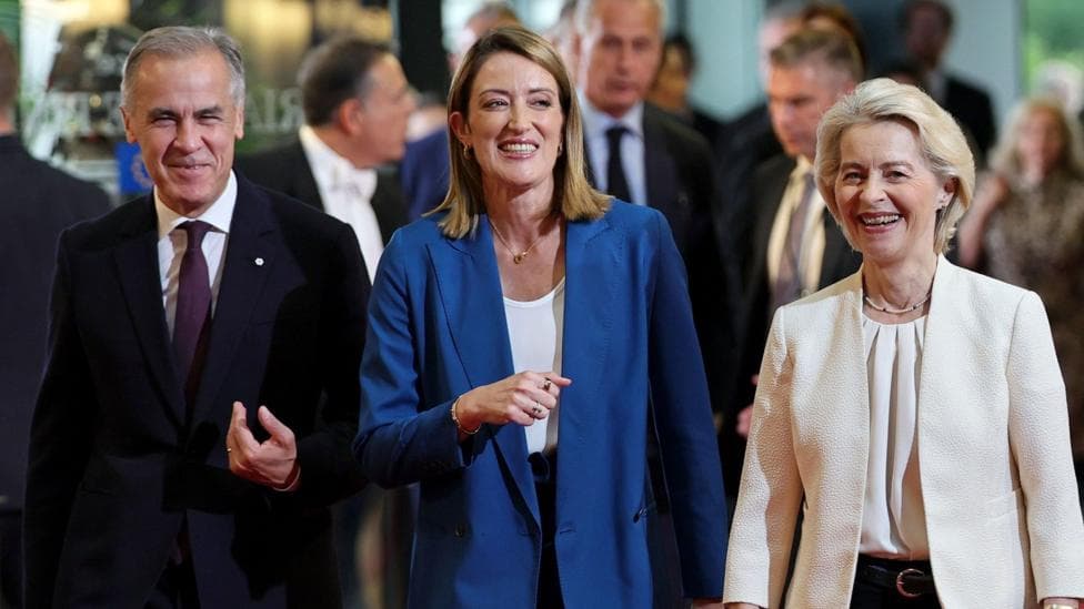 A smiling Mark Carney, wearing a suit, walks alongside European Parliament President Roberta Metsola and Ursula von der Leyen at the European Parliament