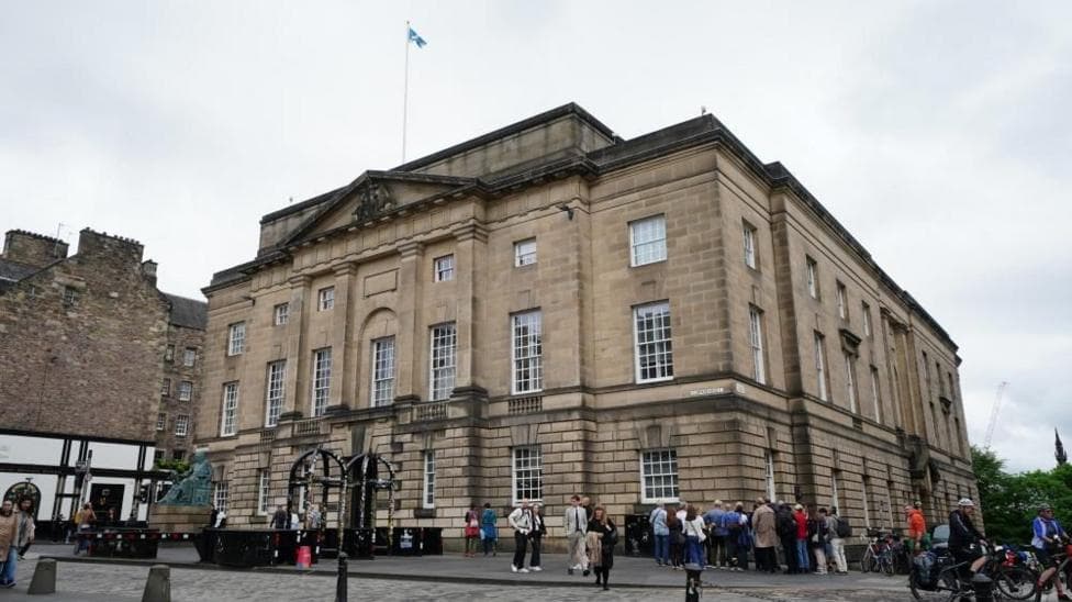 The Edinburgh High Court viewed from a street corner under an overcast sky. A Scottish flag flies from the roof, while small groups of people gather outside near the entrance.