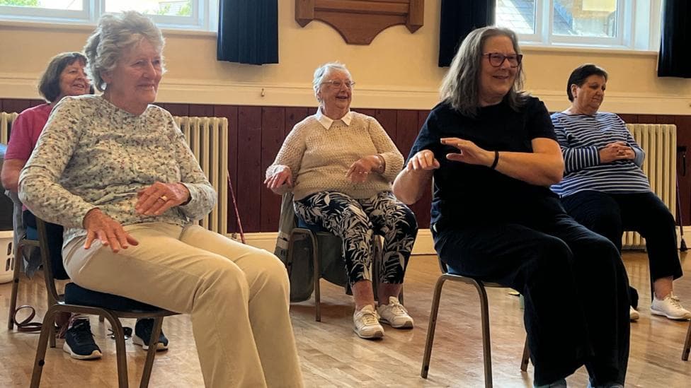 Participants sit on chairs in a community hall taking part in a seated exercise session. Several people are positioned in a circle, moving their arms while seated on a wooden floor. Large windows and wall-mounted plaques are visible in the background.