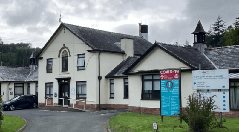 External picture of Llanidloes War Memorial Hospital. It's a cream coloured building with black window frames. A sign is seen on the right hand side. A dark coloured car is parked on the left. Trees are seen in the background.