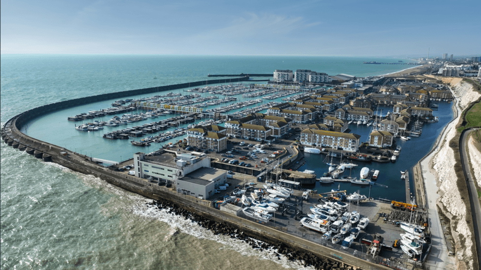 An aerial view of Brighton Marina shows curved harbour walls enclosing multiple pontoons filled with boats and yachts. Apartment blocks, marina buildings, roads and parking areas surround the basin. The sea extends into the distance alongside the Brighton coastline, with chalk cliffs visible on the right-hand side.