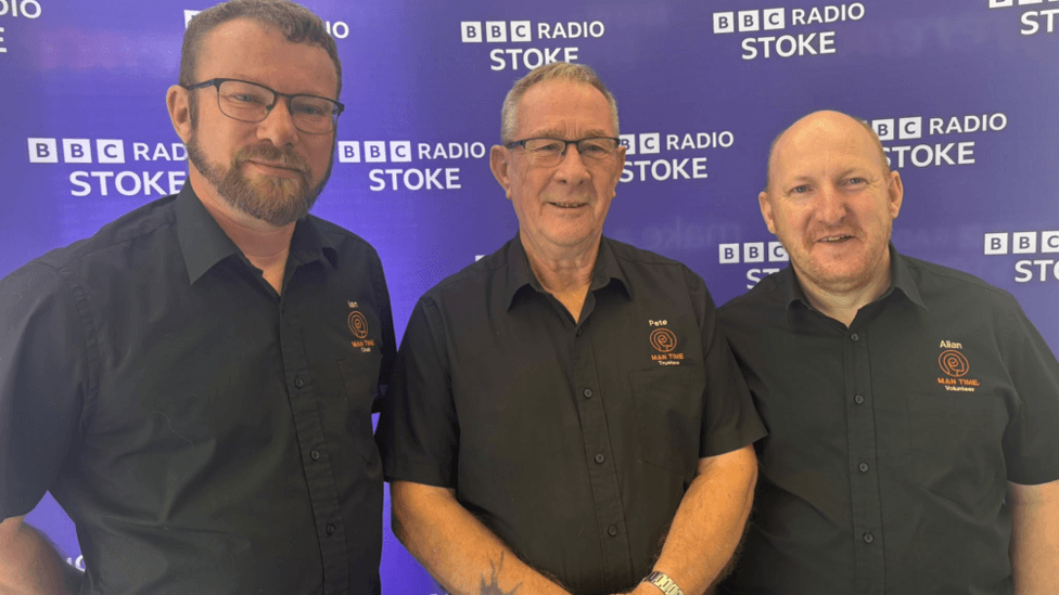 The three men stand next to each smiling at the camera. They all have black shirts on with the group's emblem on and their first names. Ian has brown hair and a beard and glasses.