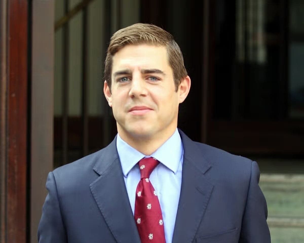 In a dark blue suit, light blue shirt and red tie, Nicholas Brandram smiles to the camera