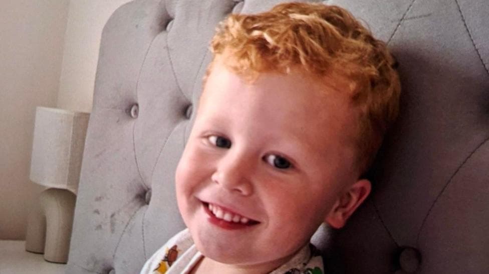 Noah, a three-year-old boy who has ginger hair. He is pictured sitting on a bed with a grey headboard and is looking directly at the camera and smiling.