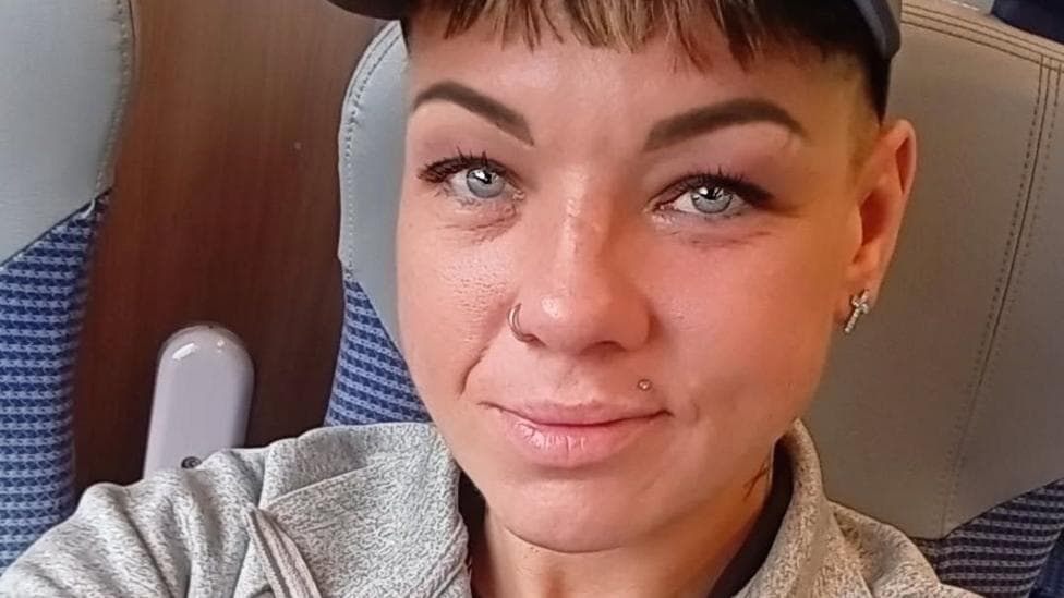 Selfie of a Jade Smith seated on a train, wearing a dark baseball cap and a light grey zip-up fleece jacket with a Berghaus logo. She has facial piercings and short hair visible beneath the cap. Blue-patterned train seats and wood-effect wall panels are visible in the background.