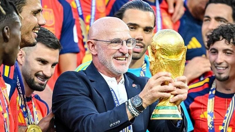 Luis de la Fuente, wearing a blue blazer, holds the gold World Cup trophy in two hands, surrounded by Spain players