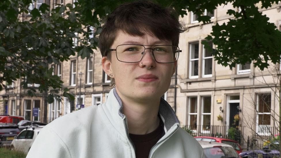 Lara wearing glasses and a light-coloured zip-up fleece standing outdoors beneath leafy trees, with stone terraced buildings and parked cars visible in the background.