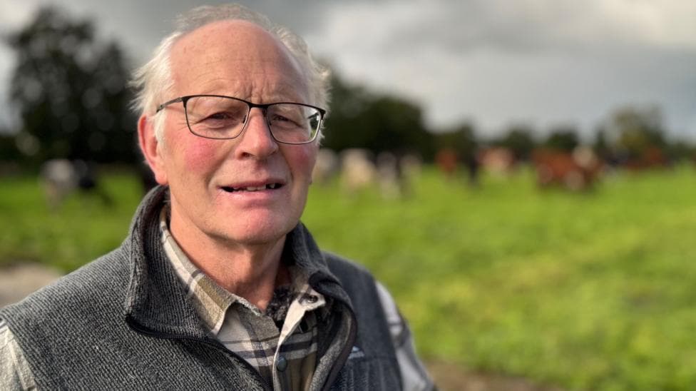 Mervyn Kelso is standing in front of a green field - there are blurred animals in the background. He is squinting at the camera. He has grey wispy hair with more on the sides than on top. Mervyn is wearing a brown checked shirt with a gilet.