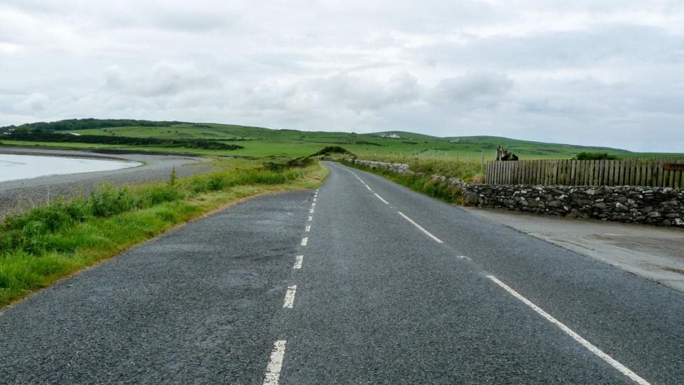 A rural road through green countryside with water to its left