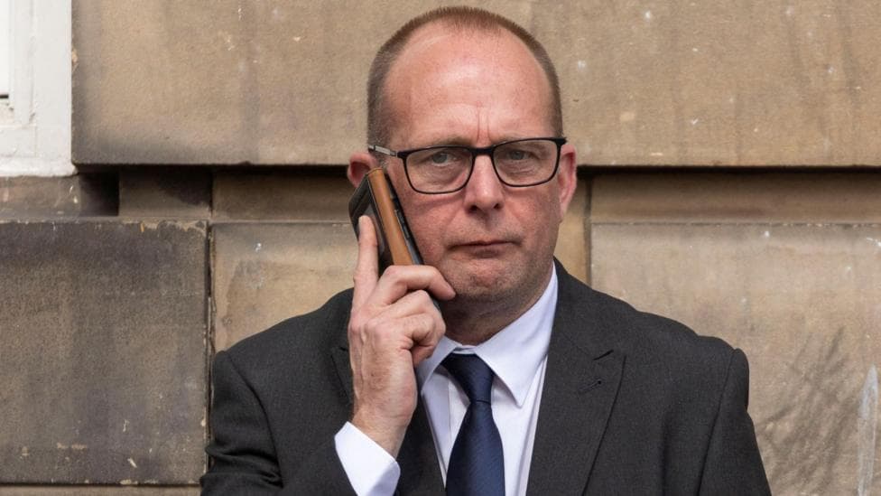 David Brown has close-cropped, dark hair. He is wearing glasses, a dark suit and tie and a white shirt. He is on a mobile phone outside a court.