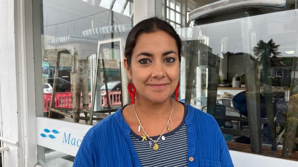 A woman wearing a stripy blue top and a bright blue shirt with earings with red lobsters on and shell necklace standing in front of her restaurant.