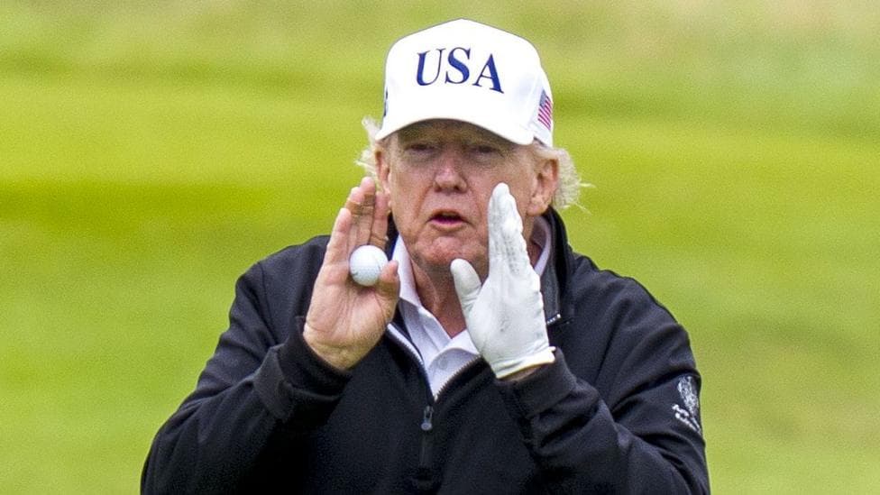 Donald Trump standing on a golf course. He has his hands raised towards his mouth, with a golf ball in one of them and a white glove on the other. He is wearing a white USA hat, and is wearing otherwise dark clothing.