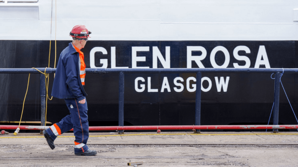 A worker wearing a red hard hat, high-visibility orange clothing and a navy jacket walks along a dockside in front of the vessel Glen Rosa. The words "GLEN ROSA" and "GLASGOW" are visible on the ship's hull in the background.