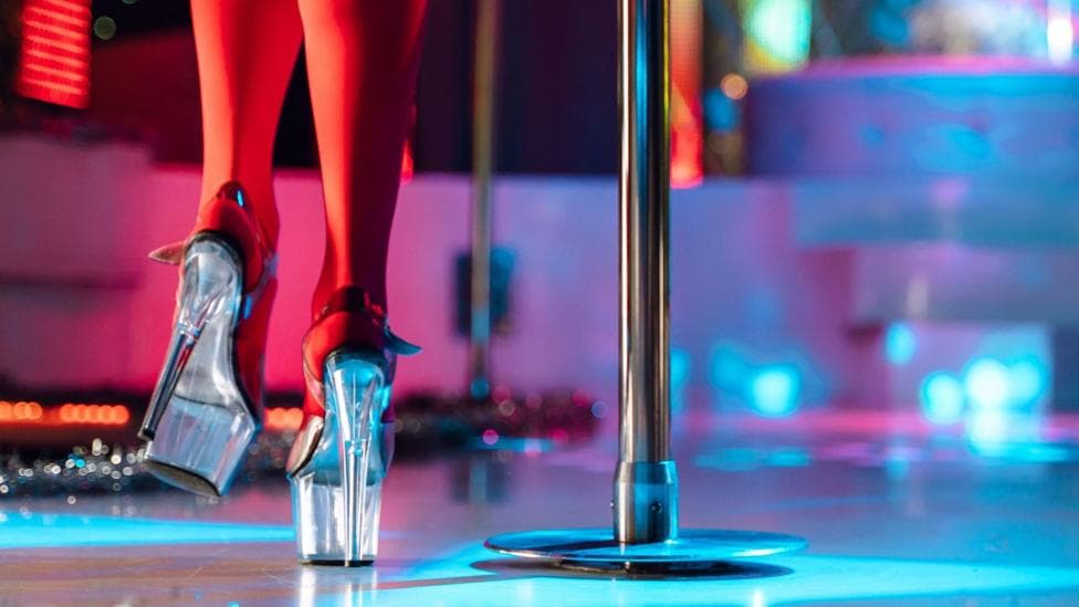 Close-up of a performer wearing clear platform high-heeled shoes walking beside a dance pole on a brightly lit stage, illuminated by vivid red, blue and pink nightclub lighting.