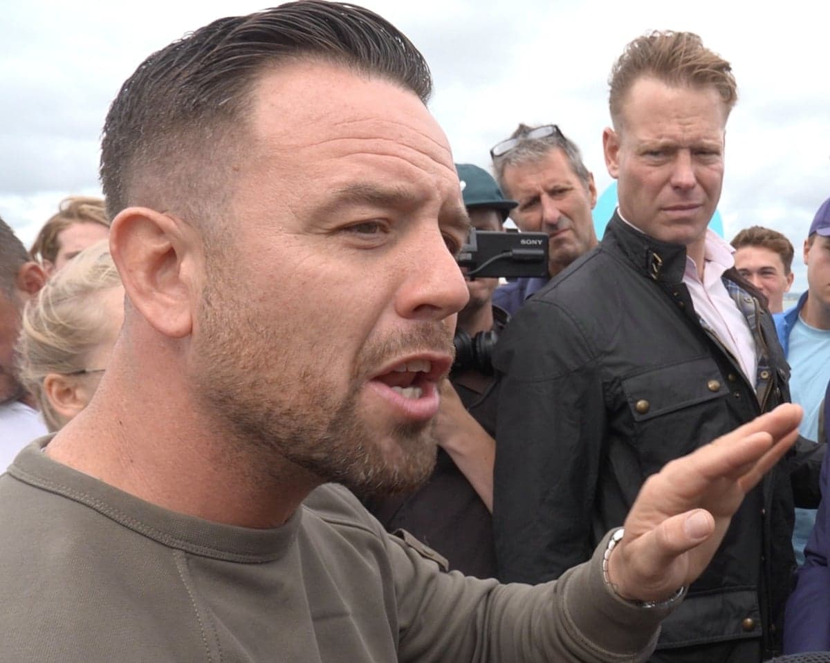 Daniel Thomas speaks in a crowd. He has short brown hair and beard and wears a light brown sweatshirt. He looks a little angry and animated with one hand raised.