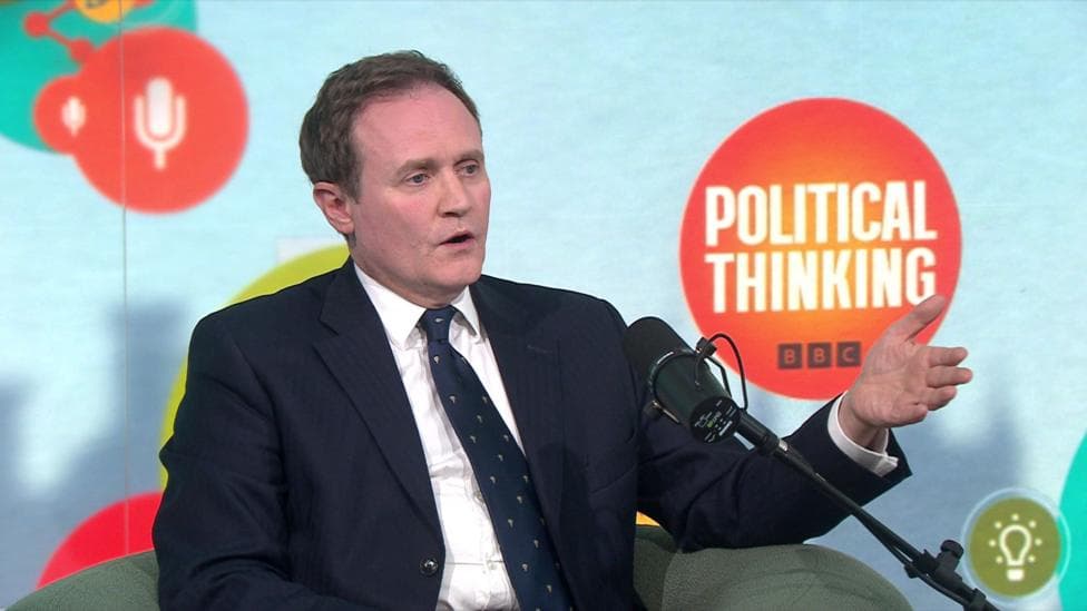 Tom Tugendhat gestures as he sits on a green chair in front of a blue background with the 'Political Thinking' logo in an orange bubble.