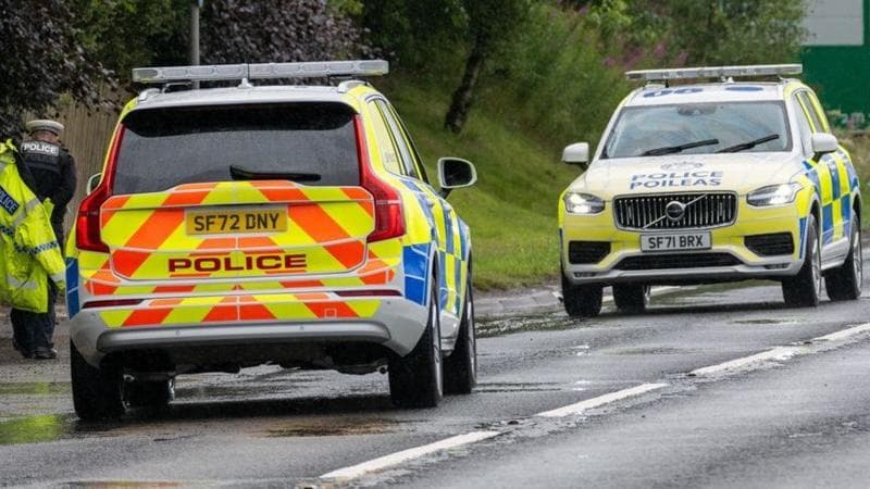 Two police cars face top face on a roadway with an officer putting on a yellow jacket beside them
