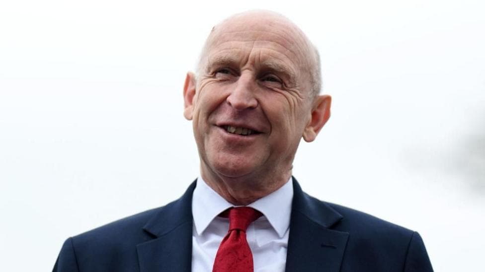 John Healey smiles while wearing a suit and red tie.